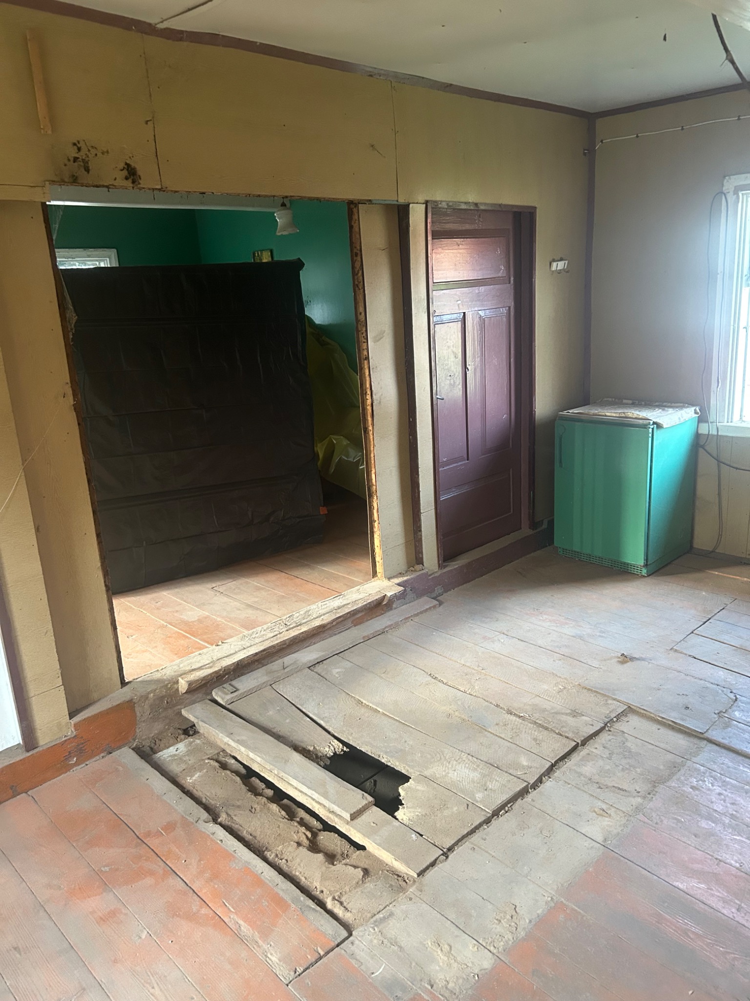 Wnętrze domu w trakcie rozbiórki: zerwana podłoga, widoczne ślady demontażu ściany działowej, zabezpieczone folią pomieszczenie obok. Stary, zielony lodówka stoi przy ścianie.