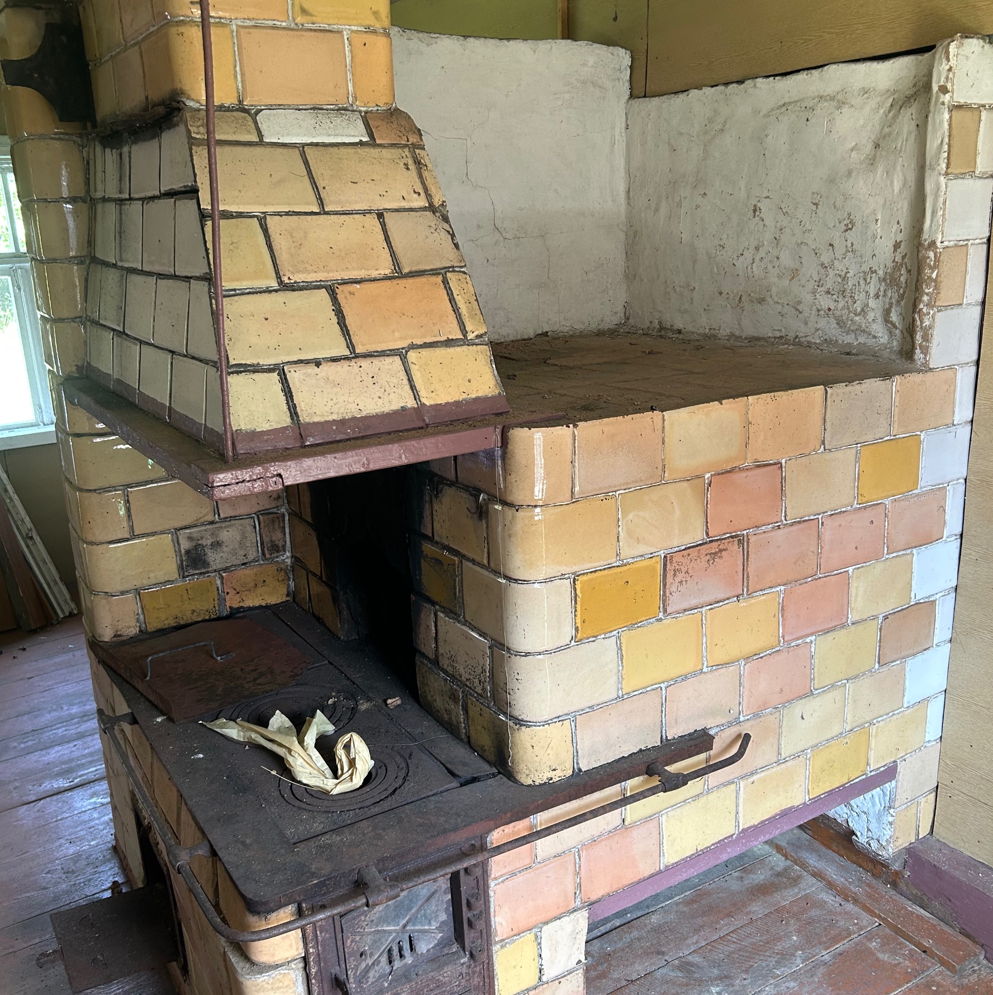 Stary piec kaflowy w odcieniach żółci i brązu, z widocznymi śladami użytkowania i napraw. Fragment ściany z ubytkami tynku. Widoczne drewniane elementy konstrukcyjne budynku.