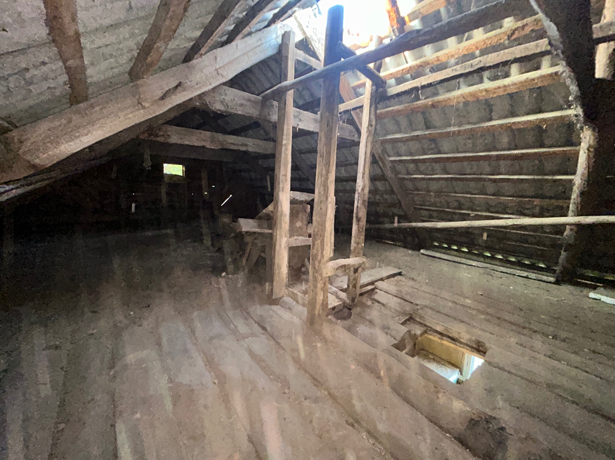 Stare, drewniane poddasze z widoczną konstrukcją dachu i podłogi. Fragmenty desek podłogowych są uszkodzone z dziurami. Widoczne drewniane podpory i niewielkie okno.