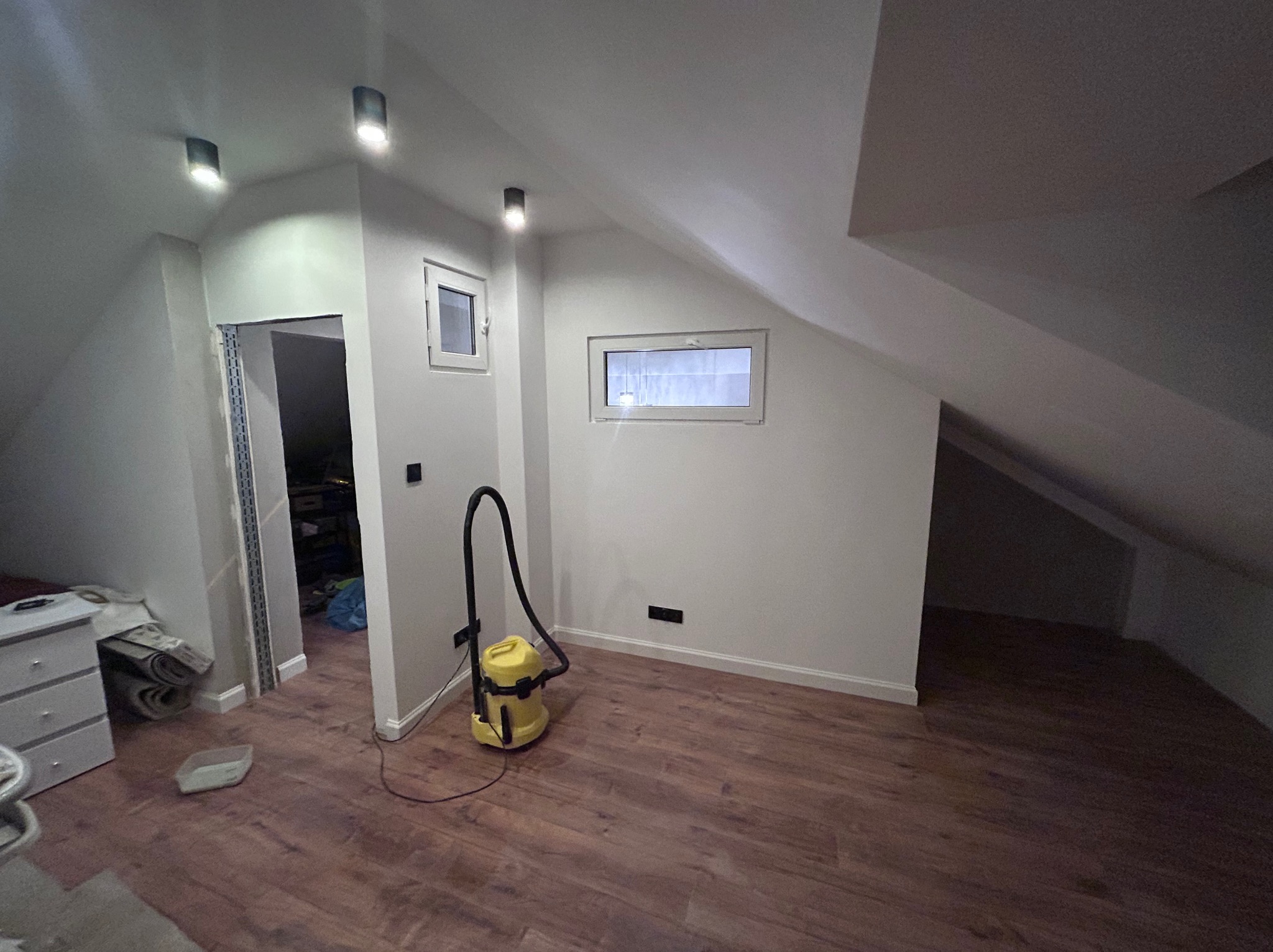 Wnętrze poddasza po remoncie: świeżo pomalowane ściany na biało, drewnopodobna podłoga, okna dachowe, widoczny odkurzacz przemysłowy i resztki materiałów budowlanych.