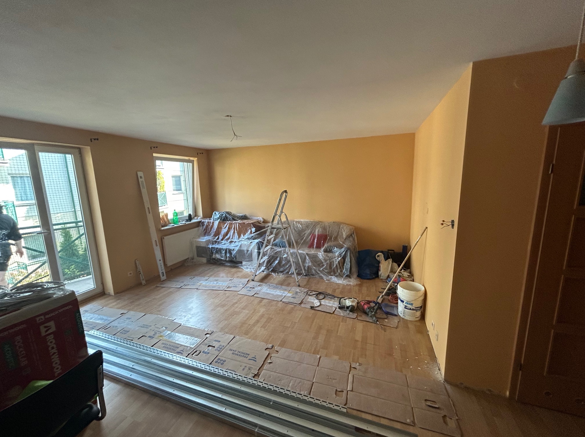 Pokój w trakcie remontu: zabezpieczone meble folią, rozłożone kartony na podłodze, widoczne narzędzia i materiały budowlane. Ściany pomalowane na żółto.