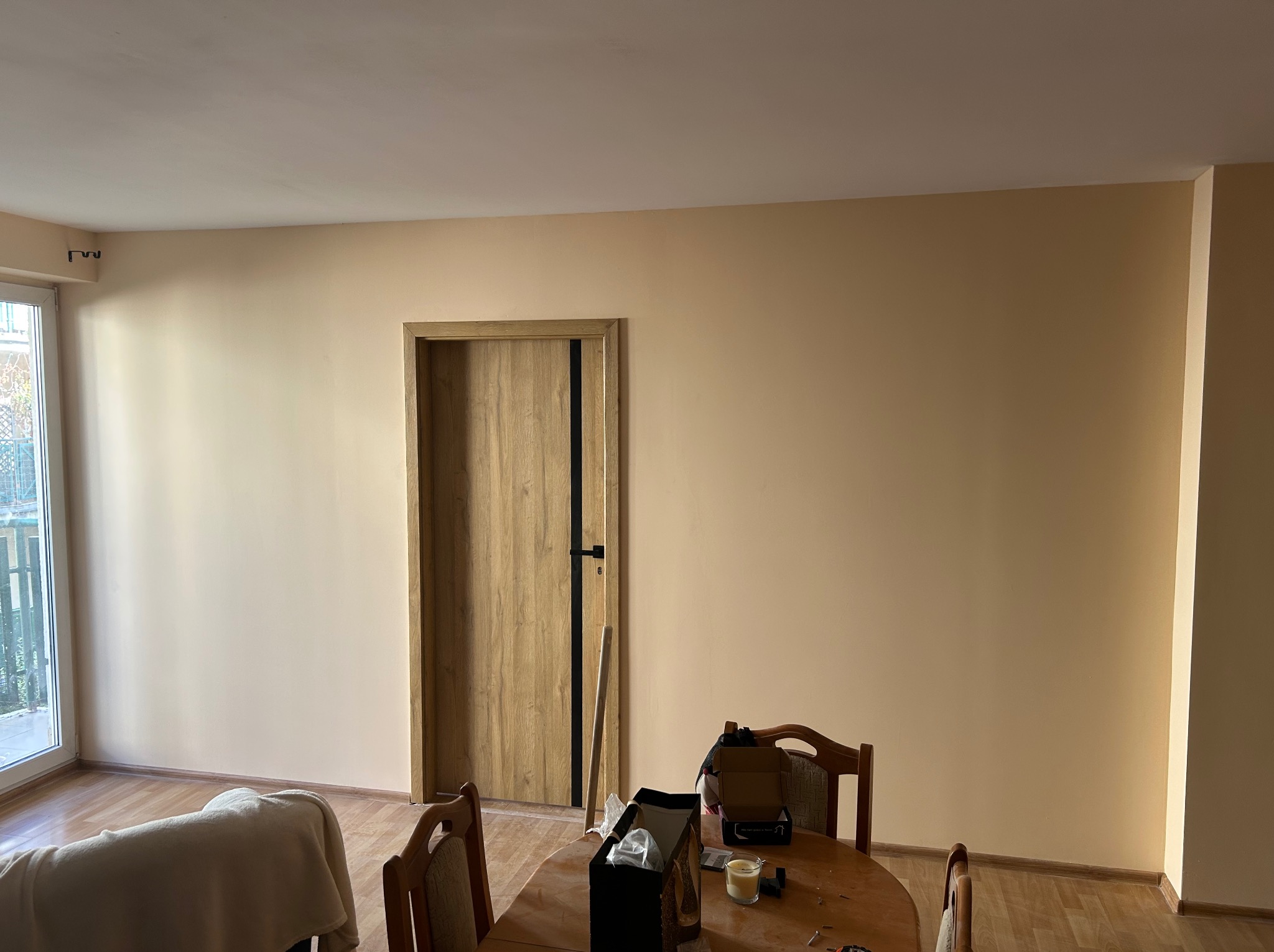 Jasne wnętrze po odświeżeniu: beżowe ściany, drewniane drzwi z czarnymi akcentami, drewniana podłoga i stół z akcesoriami malarskimi. Widok na okno.