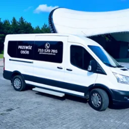 Biały bus z napisem 'Przewóz Osób' i numerem telefonu na tle nowoczesnej hali widowiskowej i zieleni. Ujęcie z boku, na pierwszym planie kostka brukowa.