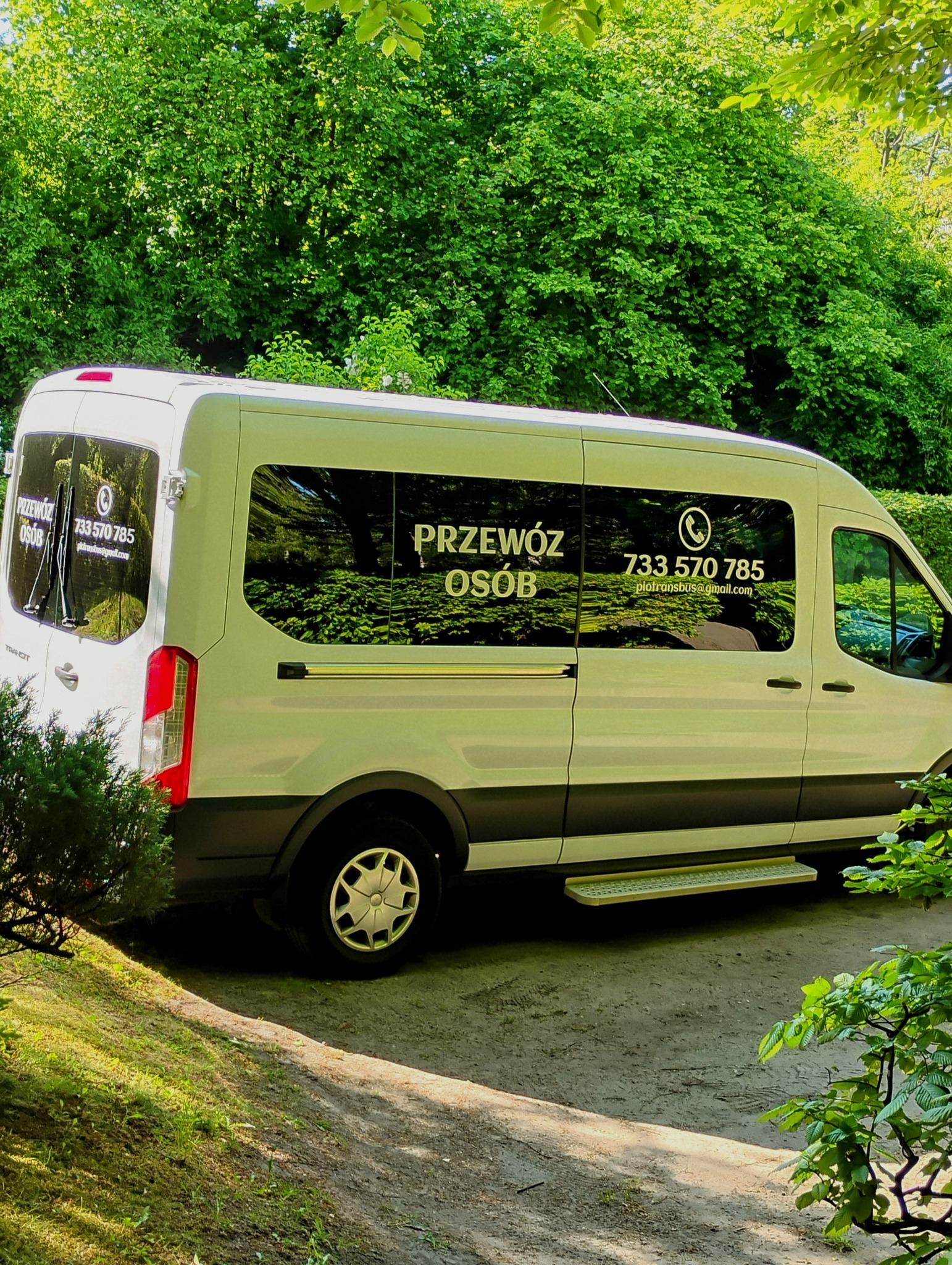 Biały bus z napisem 'Przewóz Osób' i numerem telefonu na tle zieleni. Widoczny podest ułatwiający wsiadanie. Bus zaparkowany na leśnej drodze.