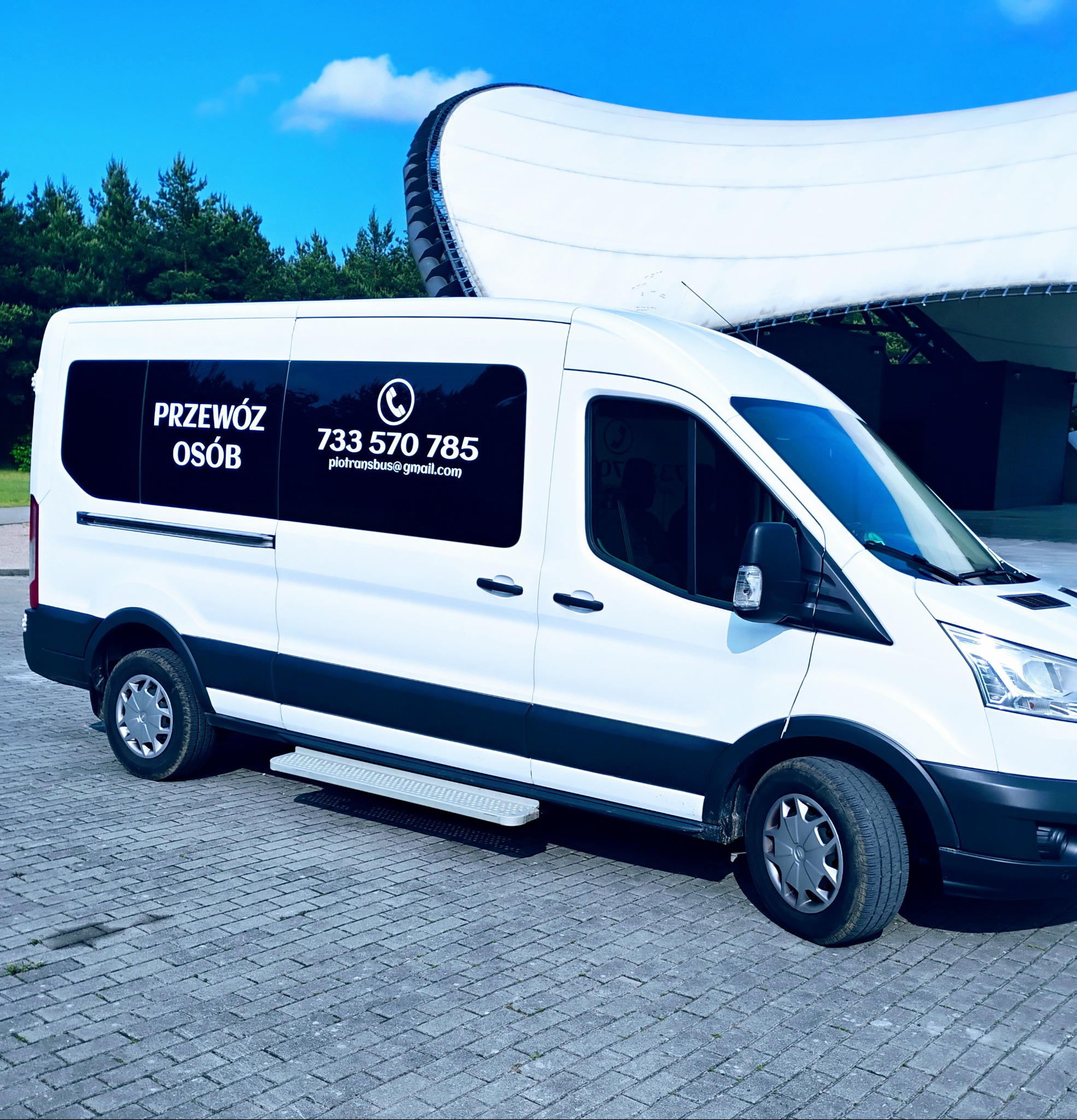 Biały bus z napisem 'Przewóz Osób' i numerem telefonu na tle nowoczesnej hali widowiskowej i zieleni. Ujęcie z boku, na pierwszym planie kostka brukowa.