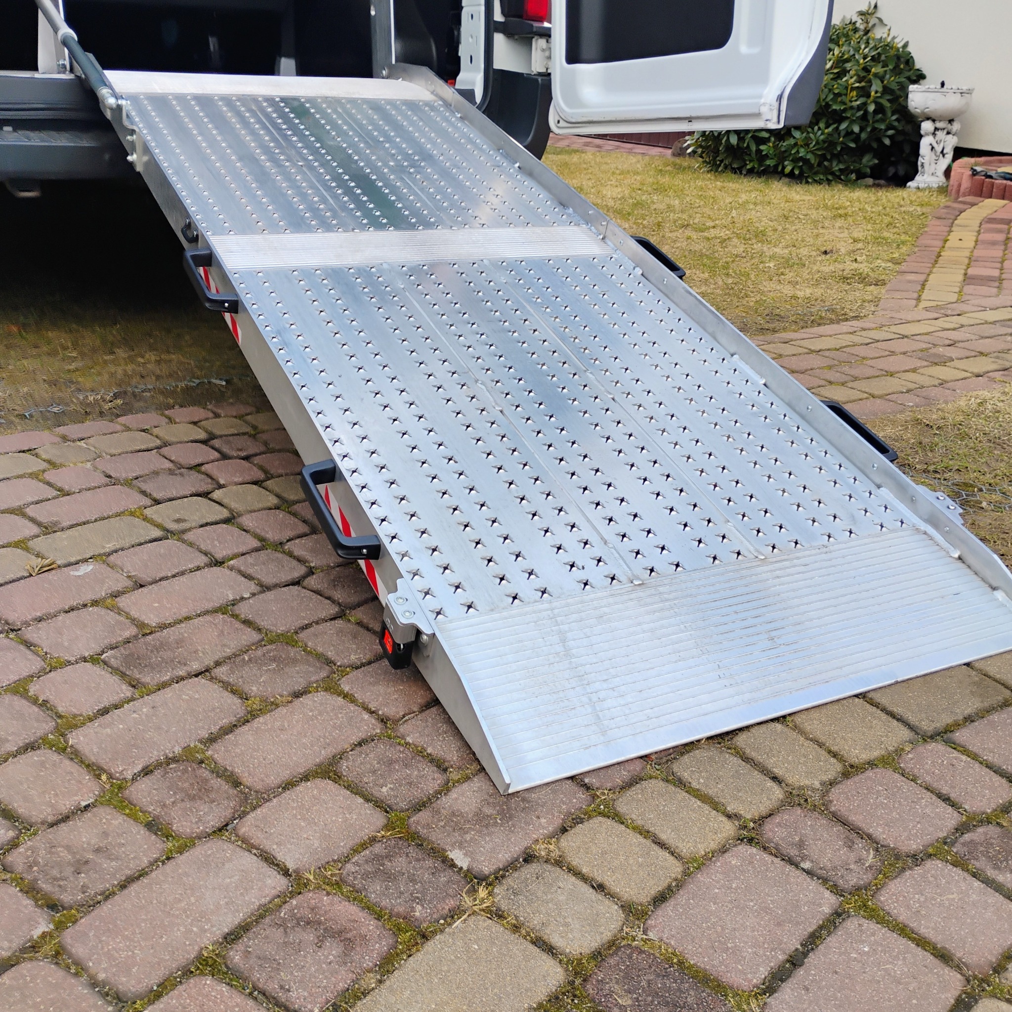 Aluminiowa rampa najazdowa do busa, ułatwiająca wjazd wózkiem inwalidzkim. Widoczna faktura antypoślizgowa i uchwyty. Tło: kostka brukowa i zieleń.
