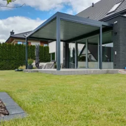 Nowoczesna, szara konstrukcja zadaszenia tarasu przylegająca do domu z dużymi oknami przesuwnymi. Zadbany trawnik i elementy wyposażenia ogrodu.