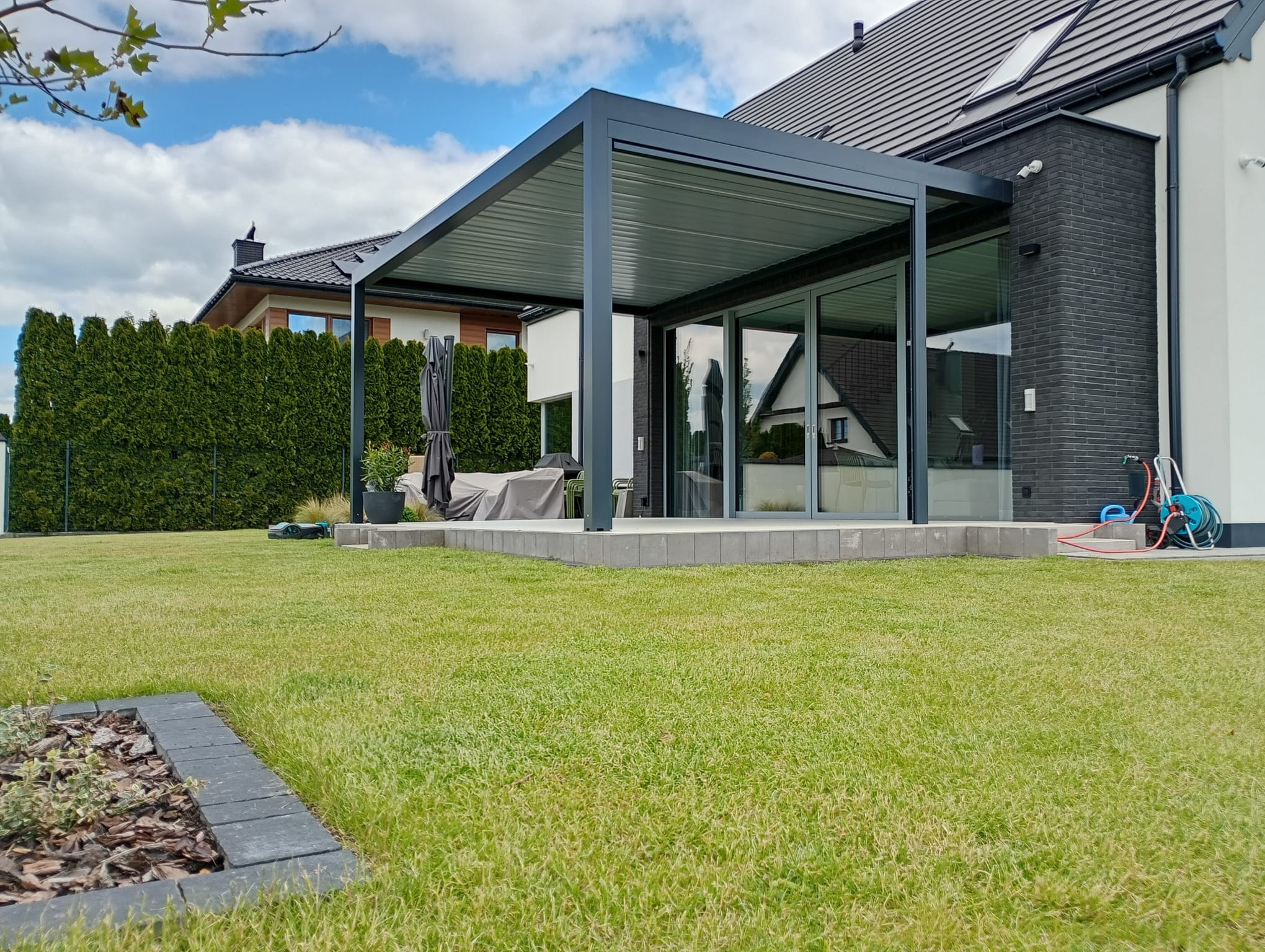 Nowoczesna, szara konstrukcja zadaszenia tarasu przylegająca do domu z dużymi oknami przesuwnymi. Zadbany trawnik i elementy wyposażenia ogrodu.