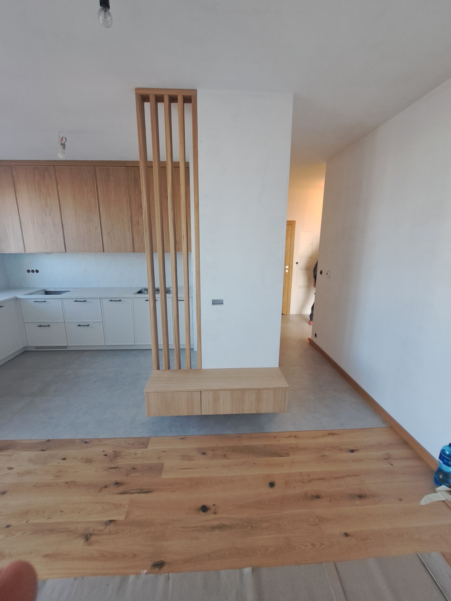 Drewniana ścianka działowa z szafką w nowoczesnym wnętrzu. Widoczna zabudowa kuchenna i parkiet. Minimalistyczny design, jasne kolory, naturalne materiały.