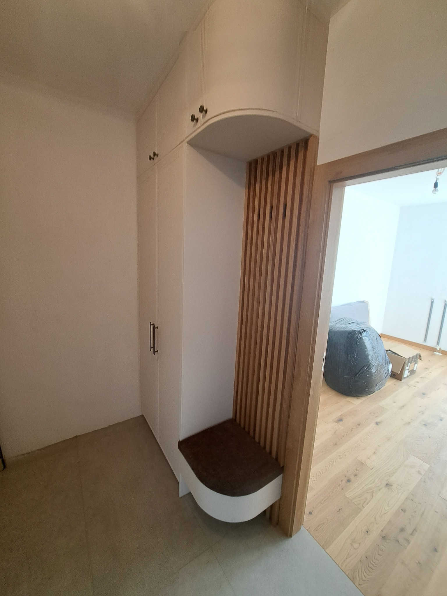 Biała szafa wnękowa z siedziskiem i drewnianą ścianką działową, widoczna z perspektywy wejścia do pomieszczenia. Minimalistyczny design, jasne wnętrze.