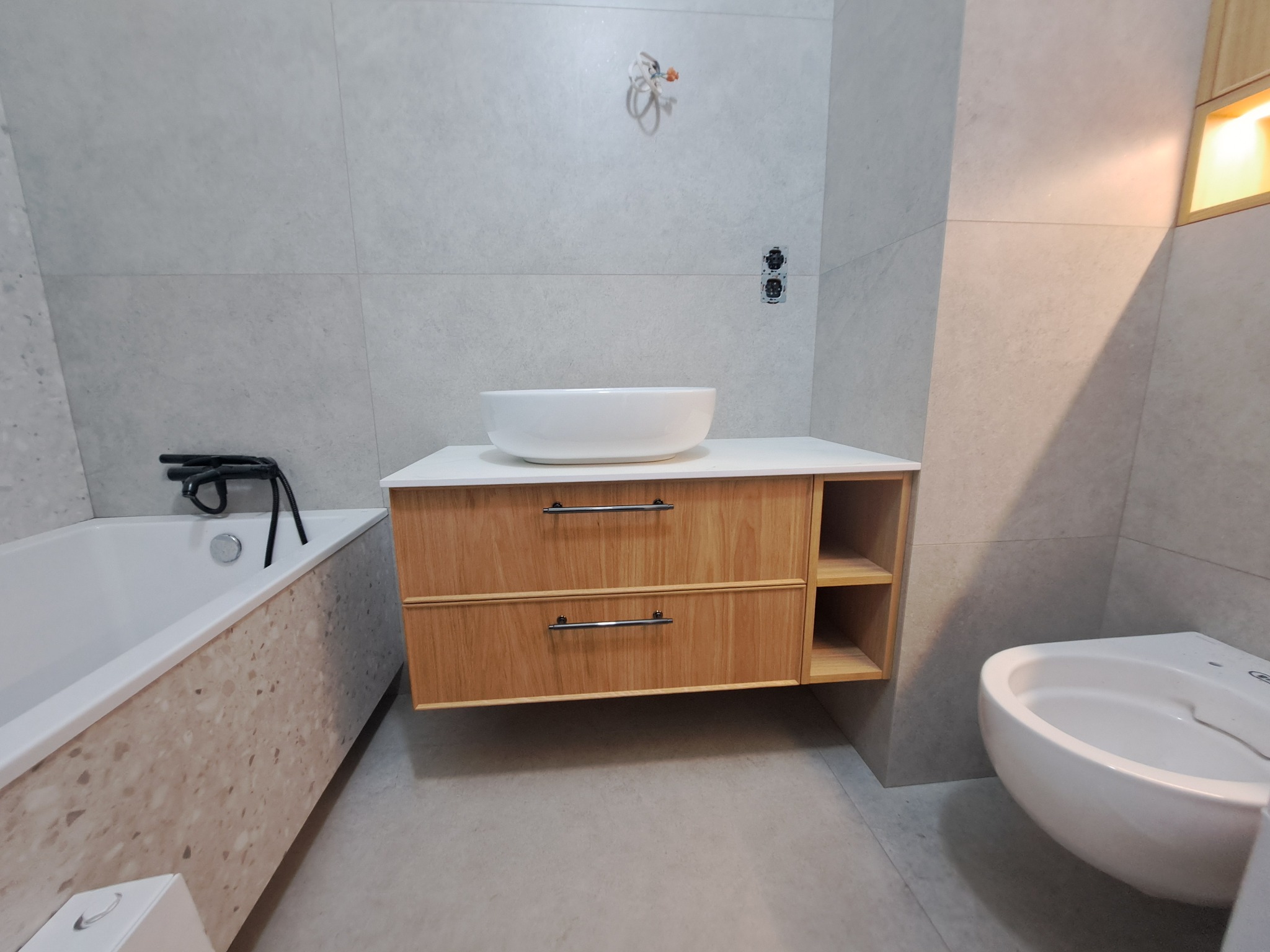 Nowoczesna łazienka z drewnianą szafką pod umywalkę, białą ceramiką i szarą płytką. Widoczne elementy instalacji elektrycznej. Minimalistyczny design.