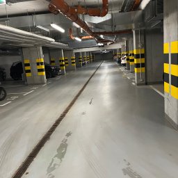 Prego Karolina Przybysz - Wnętrze parkingu podziemnego z betonowymi filarami w czarno-żółte pasy. Widoczne instalacje pod sufitem i ślady po umyciu posadzki.