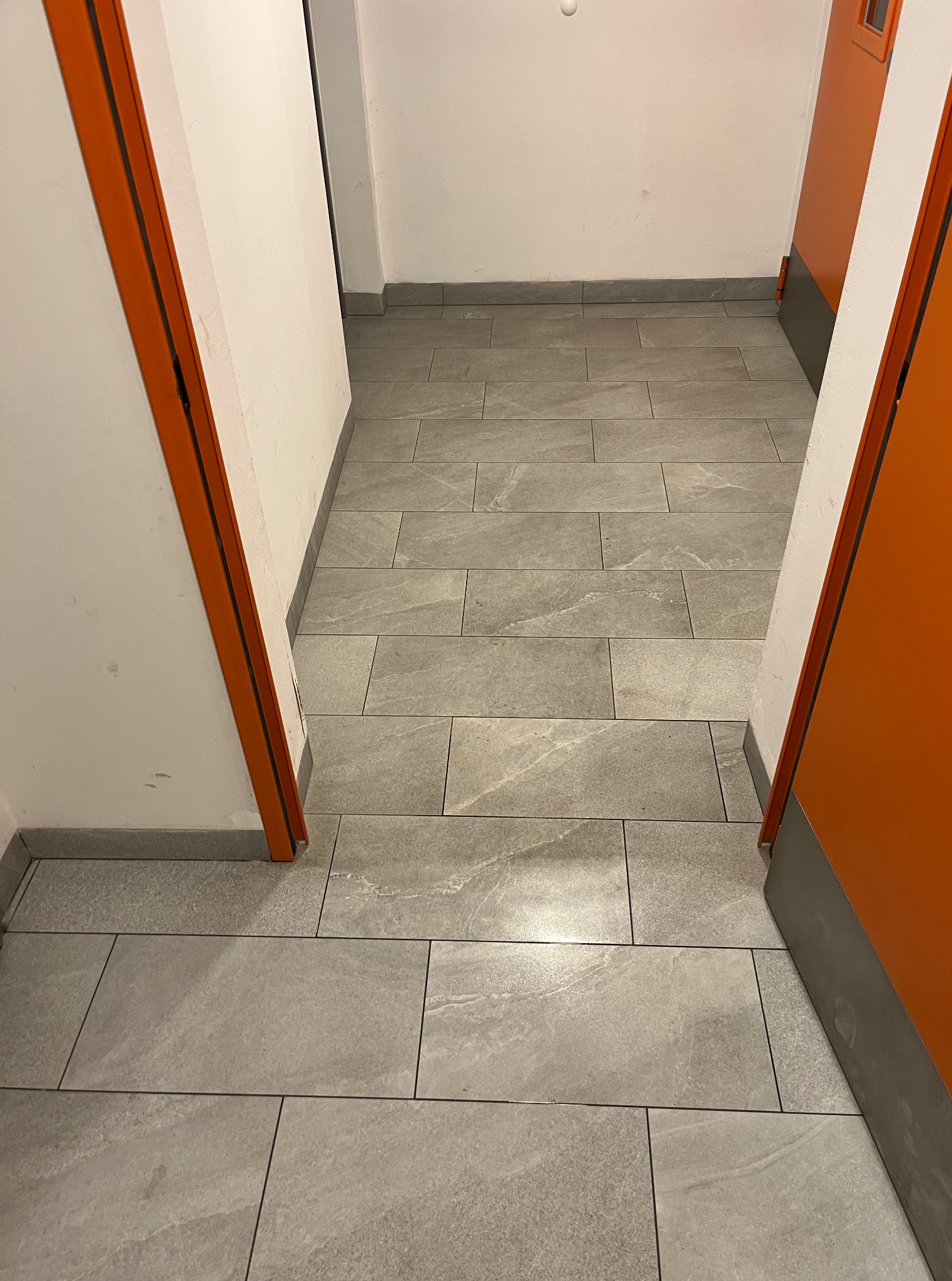 Korytarz z szarymi płytkami podłogowymi, pomalowany na biało, z pomarańczowymi drzwiami po obu stronach. Widok z góry na czystą podłogę.