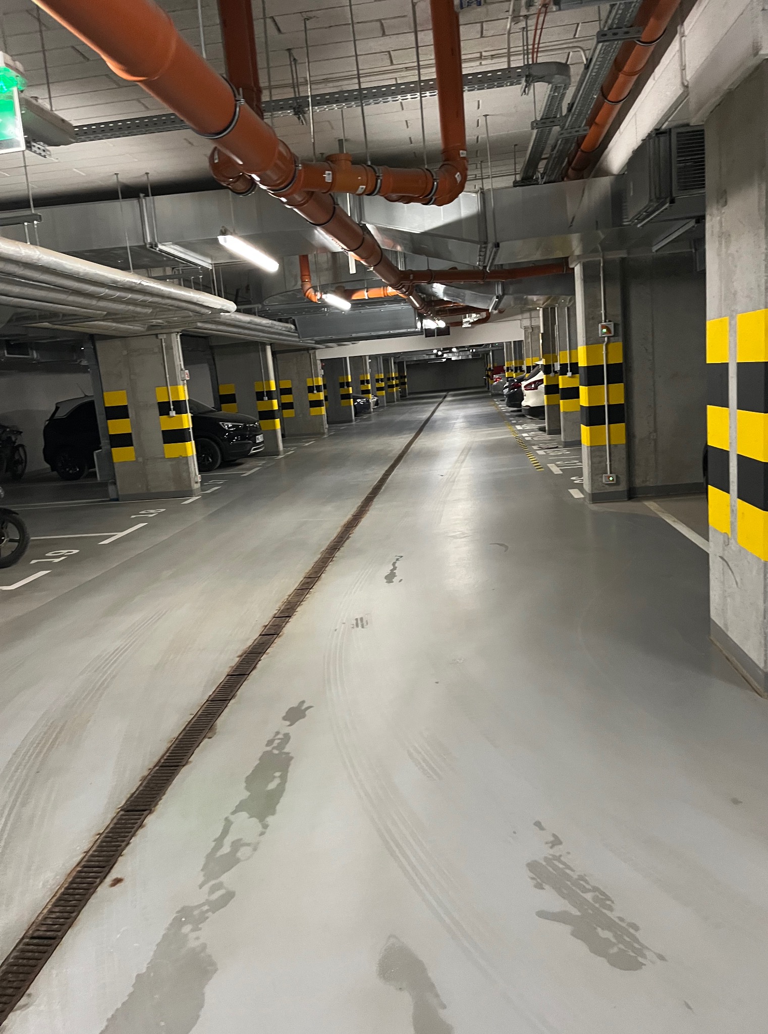 Wnętrze parkingu podziemnego z betonowymi filarami w czarno-żółte pasy. Widoczne instalacje pod sufitem i ślady po umyciu posadzki.