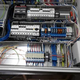 Rozdzielnica elektryczna z licznymi przewodami, przekaźnikami i zabezpieczeniami, widok z góry. Precyzyjne połączenia i uporządkowany układ komponentów.