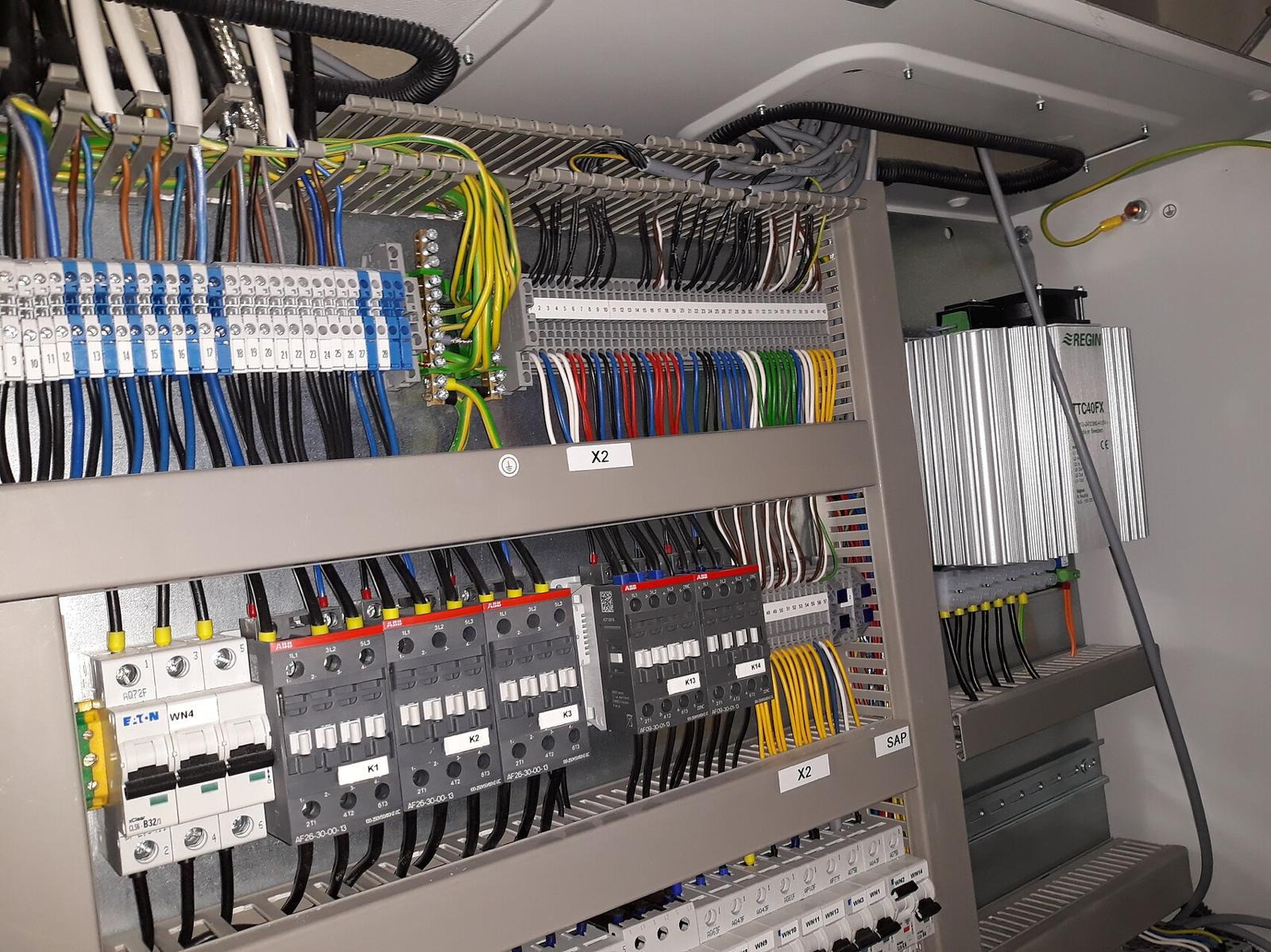 Rozdzielnica elektryczna z podłączonymi przewodami, bezpiecznikami i stycznikami. Widoczne oznaczenia X2, K1, K2, K13, K14, SAP oraz moduł Regin TC40FX.