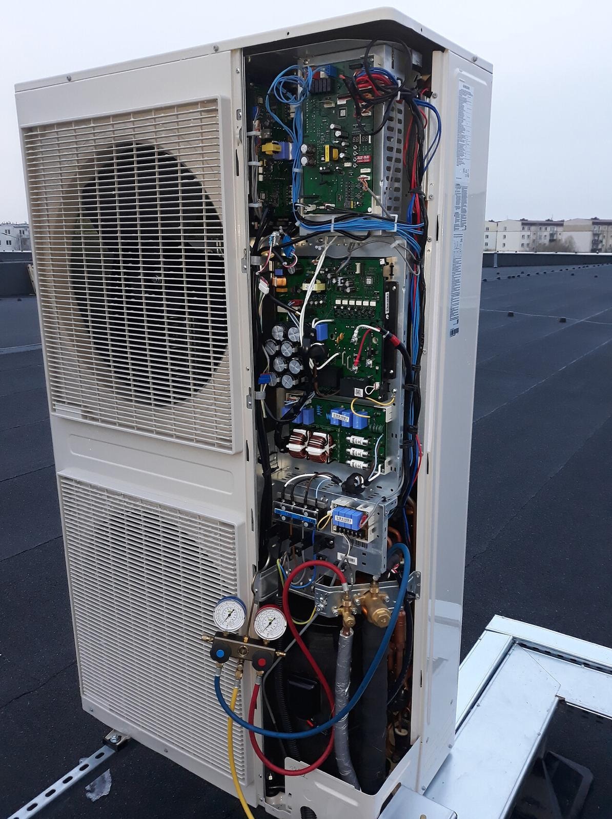 Rozłożona jednostka zewnętrzna klimatyzacji na dachu, z widocznymi podzespołami elektronicznymi, przewodami i manometrami. Widok z boku na tle zabudowań miejskich.
