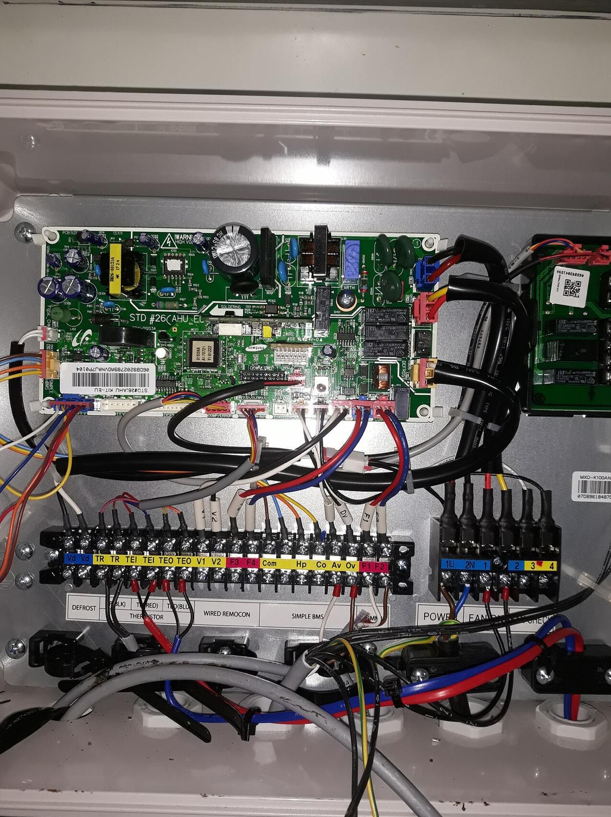 Rozdzielnica elektryczna z licznymi przewodami, płytką drukowaną i złączami. Widoczne oznaczenia i etykiety na przewodach. Skrzynka w kolorze szarym.