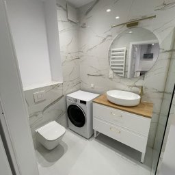 K&V SEVKO SPÓŁKA Z OGRANICZONĄ ODPOWIEDZIALNOŚCIĄ - Biała łazienka z marmurowymi płytkami, umywalką na drewnianym blacie, pralką i toaletą. Okrągłe lustro z oświetleniem. Nowoczesny design.