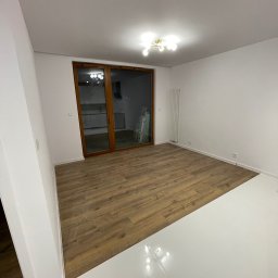 K&V SEVKO SPÓŁKA Z OGRANICZONĄ ODPOWIEDZIALNOŚCIĄ - Jasne wnętrze po remoncie w Białymstoku: panele podłogowe imitujące drewno, białe ściany i podłoga. Widoczne okno balkonowe i grzejnik.