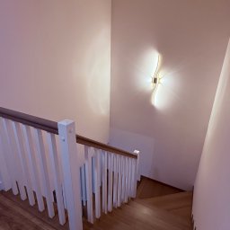 Vladimir Sevko - Drewniane schody z białymi balustradami, oświetlone nowoczesnym kinkietem na białej ścianie. Ujęcie z góry, podkreślające detale wykończenia.