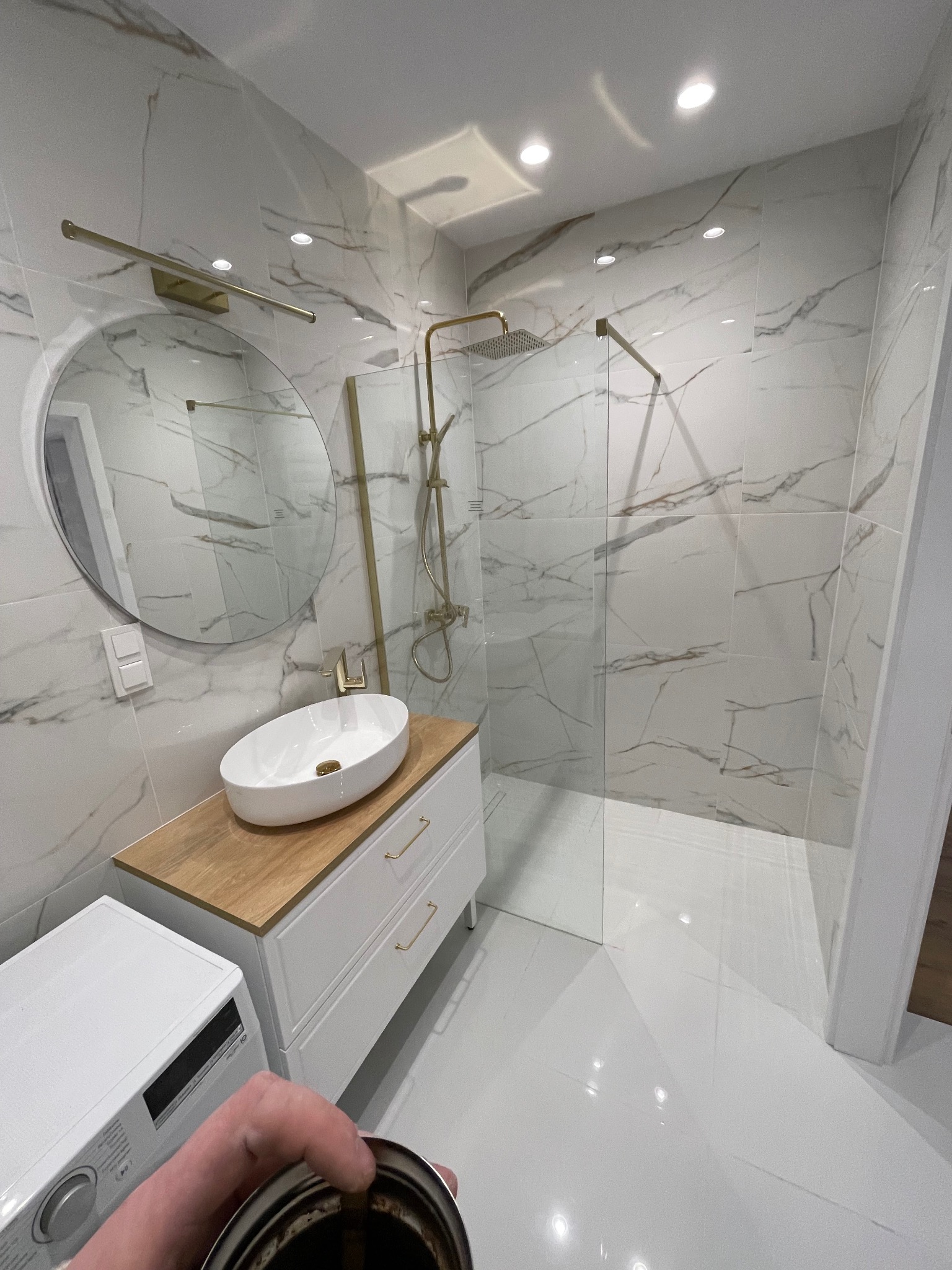 Nowoczesna łazienka z kabiną prysznicową typu walk-in, umywalką nablatową na szafce i okrągłym lustrem. Białe płytki imitujące marmur z delikatnym złotym akcentem.