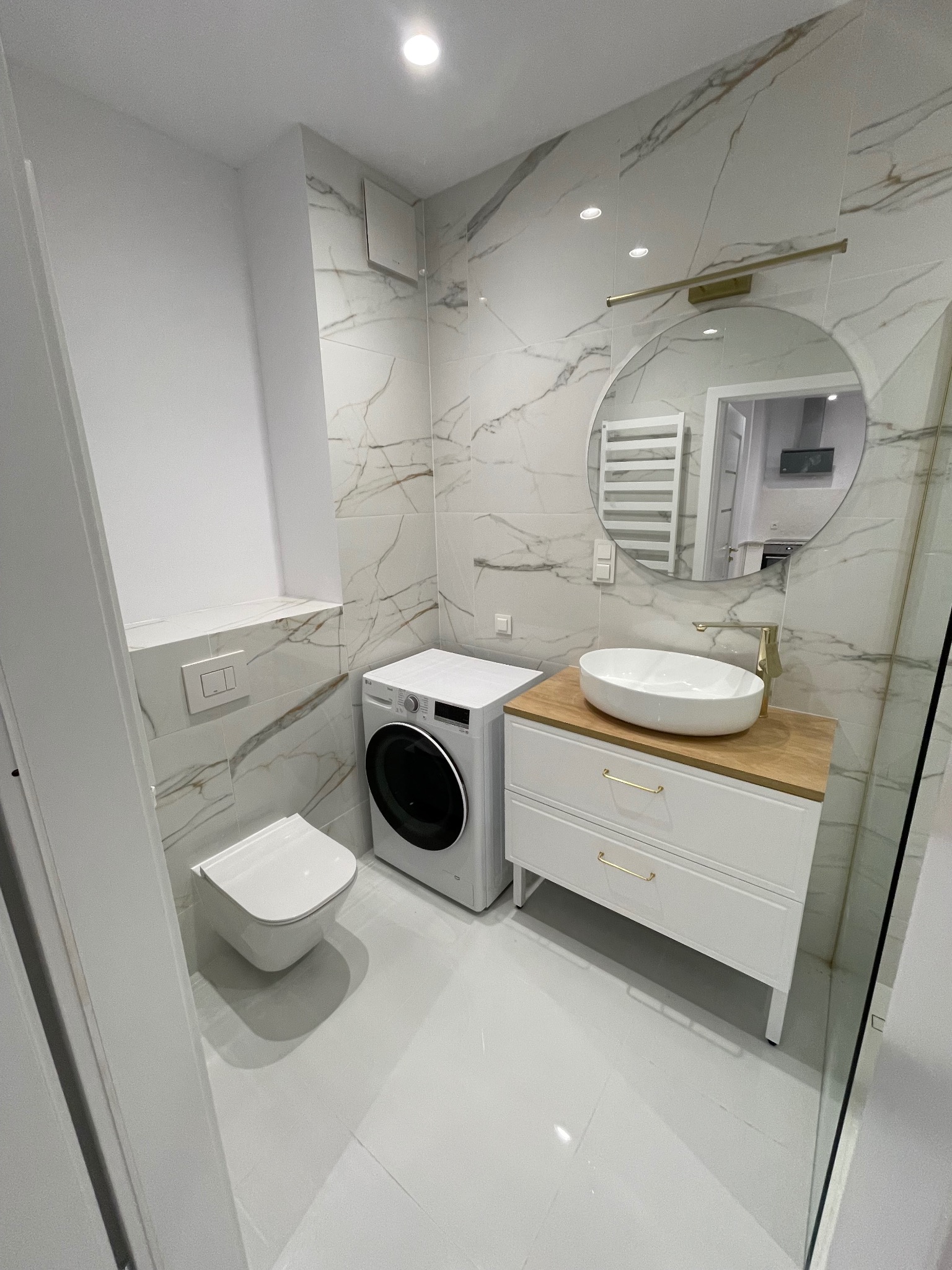 Biała łazienka z marmurowymi płytkami, umywalką na drewnianym blacie, pralką i toaletą. Okrągłe lustro z oświetleniem. Nowoczesny design.