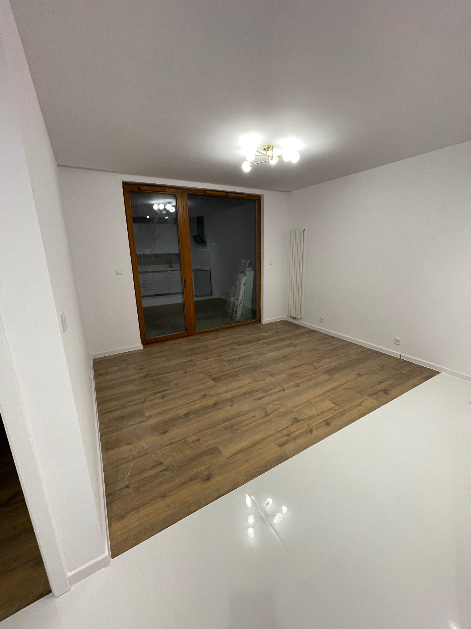 Jasne wnętrze po remoncie w Białymstoku: panele podłogowe imitujące drewno, białe ściany i podłoga. Widoczne okno balkonowe i grzejnik.