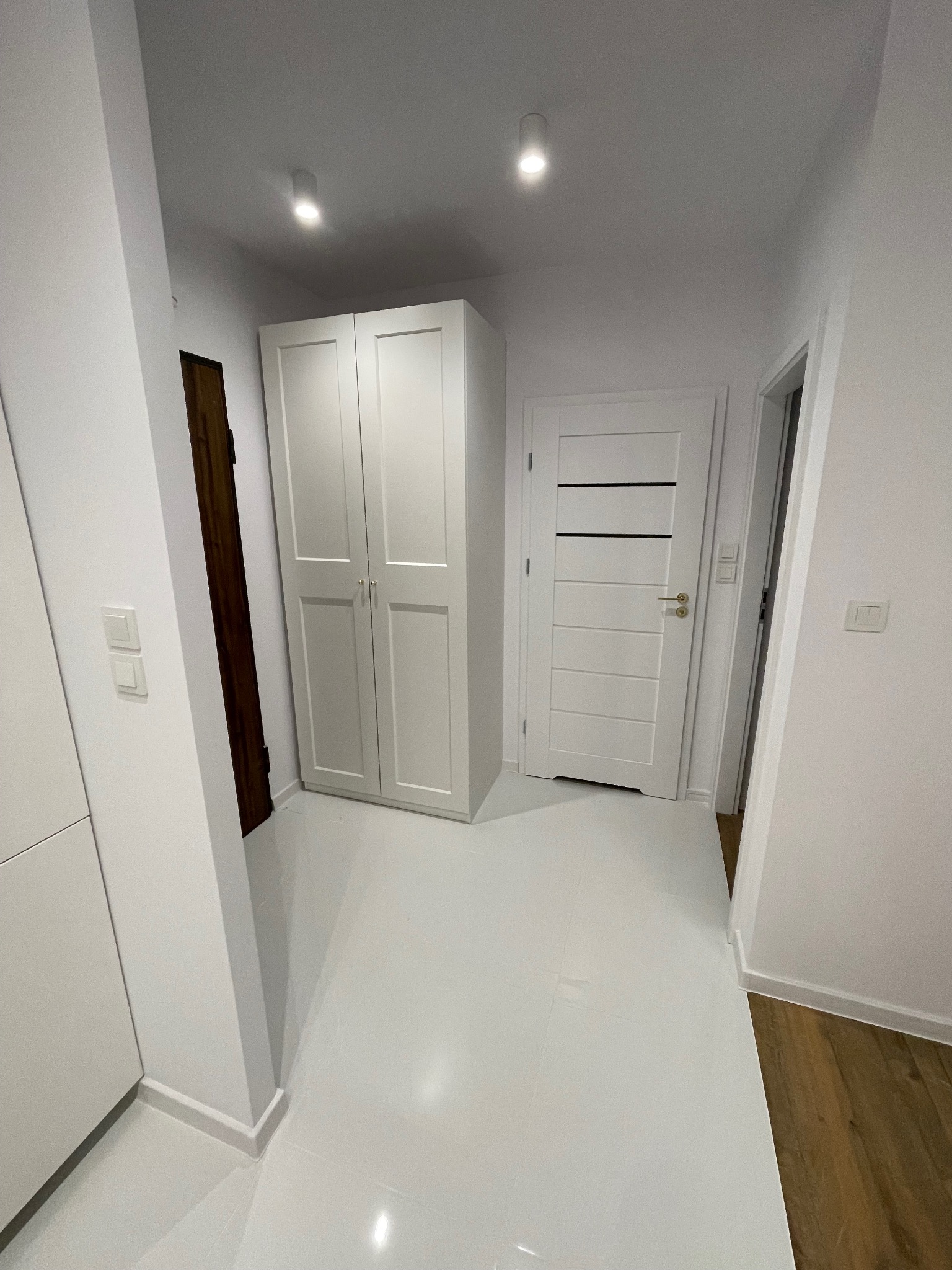 Białe wnętrze korytarza z szafą, drzwiami i błyszczącą podłogą. Widoczne detale wykończenia, takie jak listwy przypodłogowe i włączniki światła.
