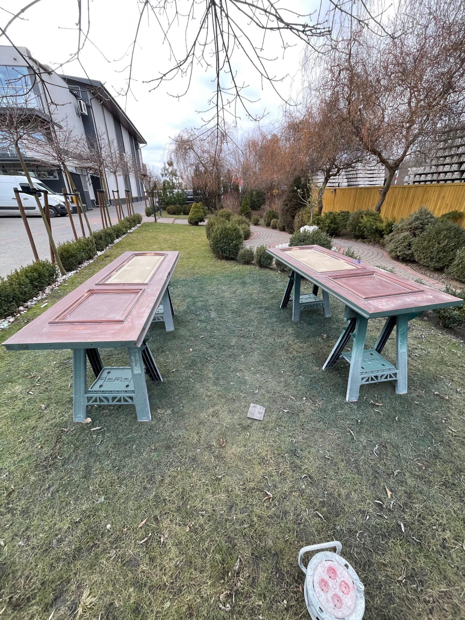 Dwie drewniane, odnowione ramy drzwiowe na kozłach malarskich, ustawione na trawniku przed budynkiem. Widoczna lampa warsztatowa.