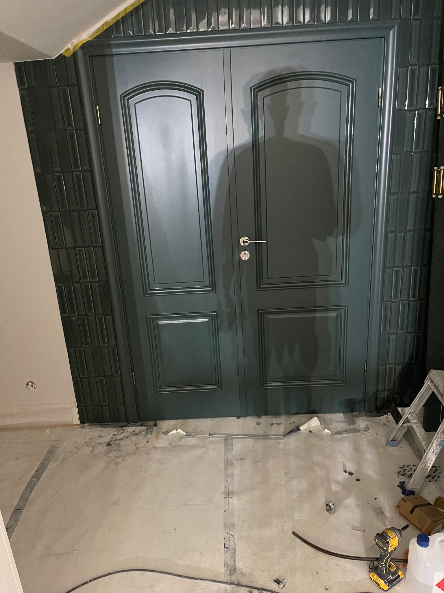 Zielone, dwuskrzydłowe drzwi z frezowanymi panelami, zamontowane w ścianie z ciemnozielonej, błyszczącej glazury typu 'metro'. Na podłodze widoczne ślady prac wykończeniowych.