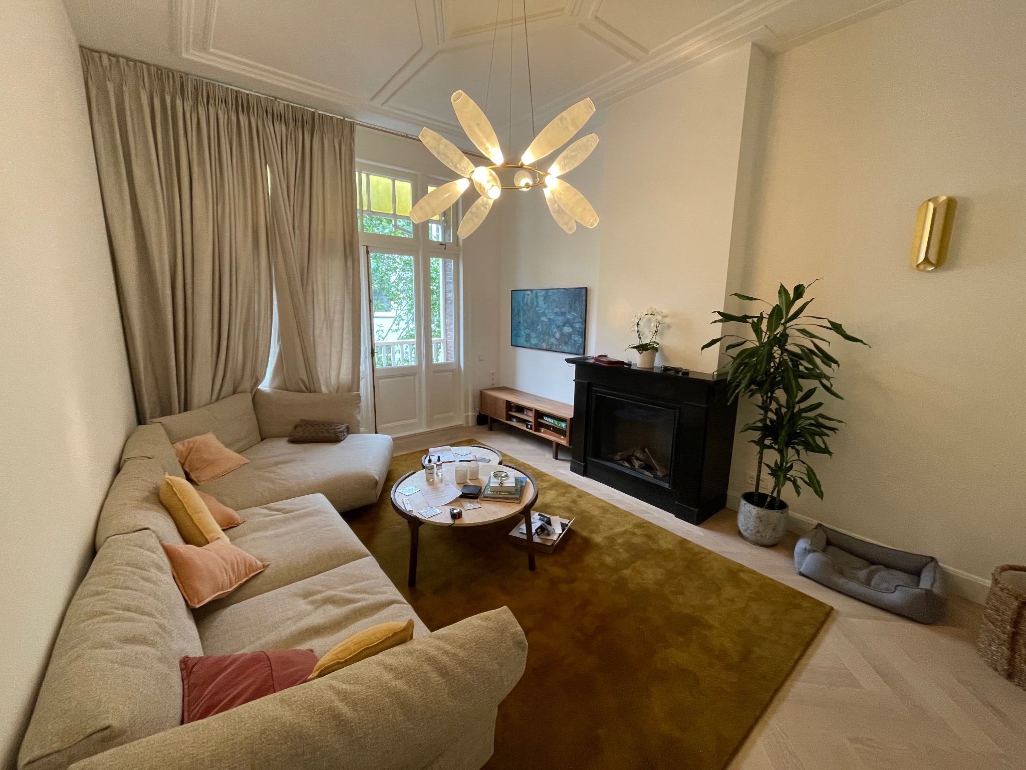 Jasny salon z beżową sofą narożną, okrągłym stolikiem kawowym i czarnym kominkiem. Na ścianie złoty kinkiet, a nad kominkiem telewizor. Całość oświetla designerski żyrandol.