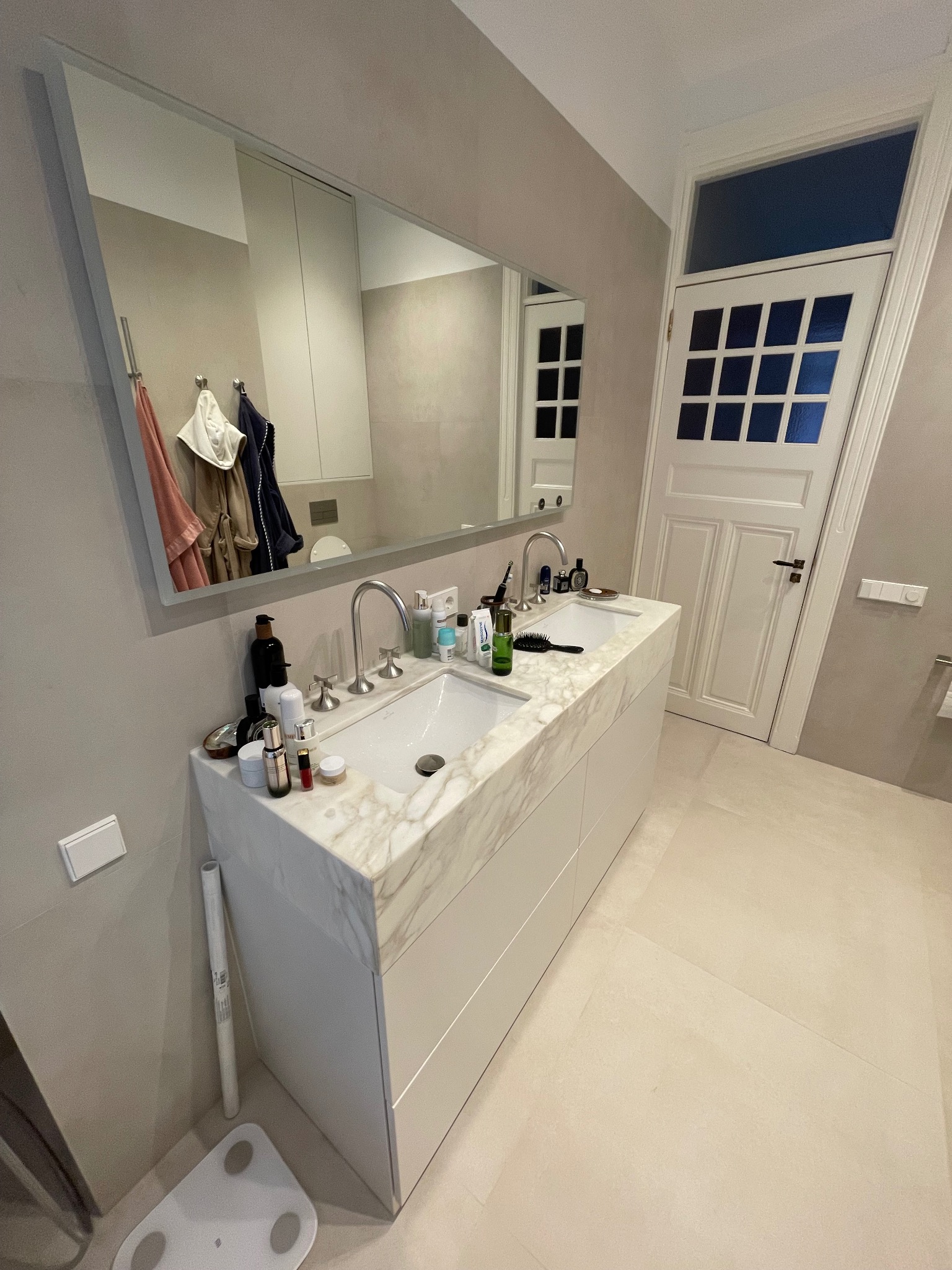 Jasna łazienka z podwójną umywalką na marmurowym blacie, duże lustro odbija fragment szafy i drzwi z szybkami. Na blacie kosmetyki i akcesoria łazienkowe.