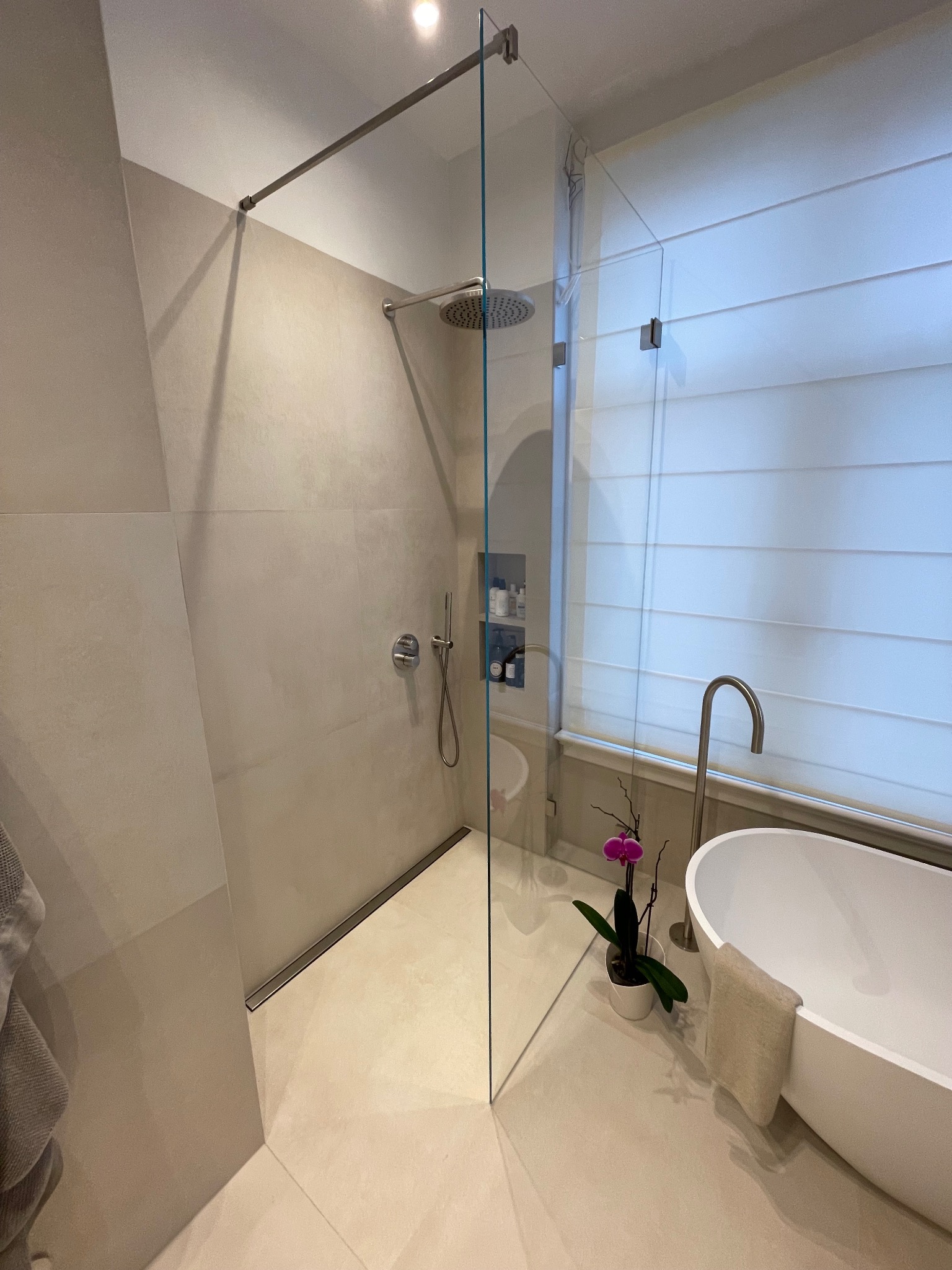Nowoczesna łazienka z kabiną typu walk-in, deszczownicą i wolnostojącą wanną. Minimalistyczny design, beżowe płytki, szklana ściana kabiny, orchidea w doniczce.