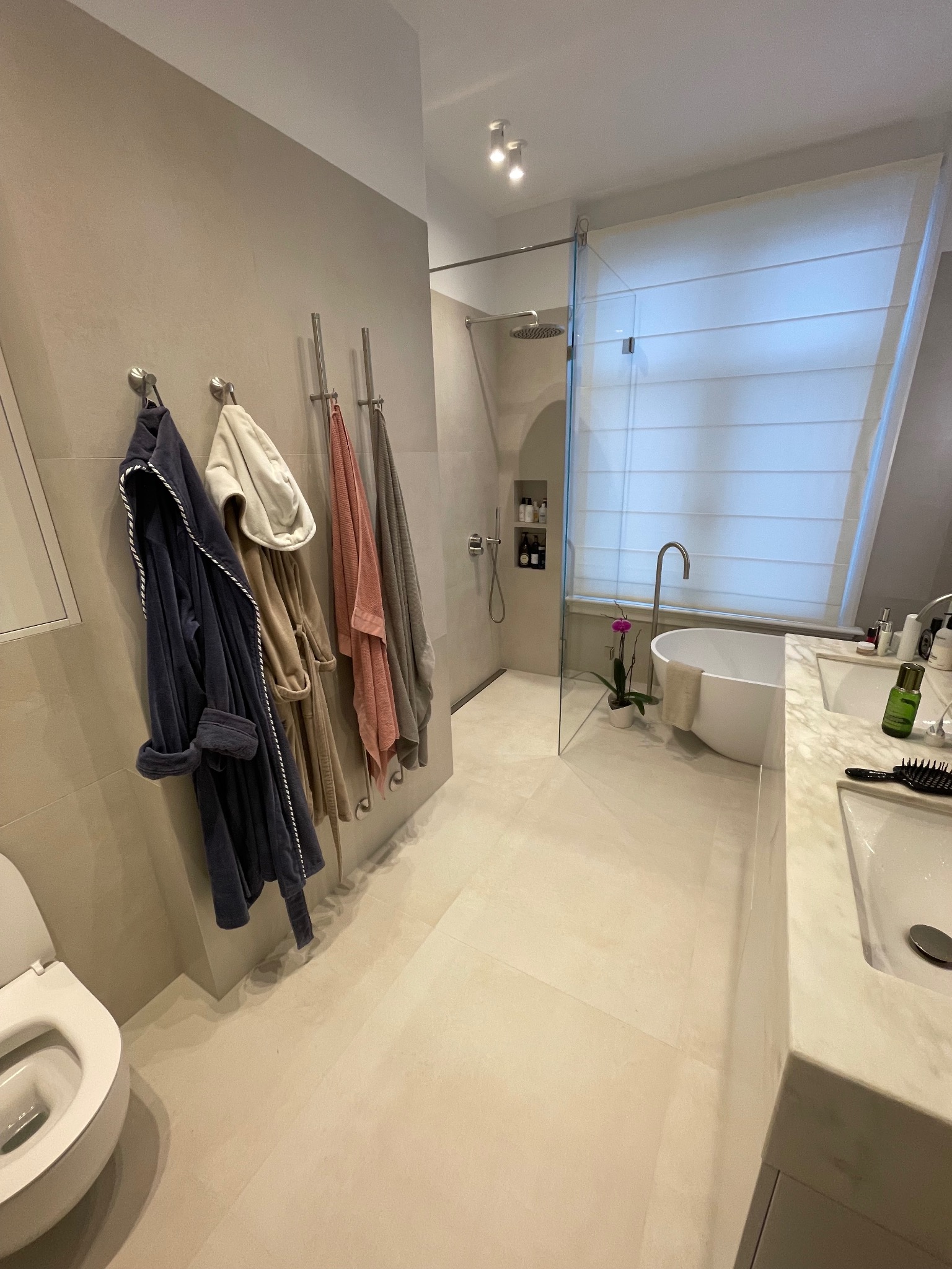 Nowoczesna łazienka z beżowymi płytkami, kabiną prysznicową typu walk-in, wanną wolnostojącą i ręcznikami na wieszakach. Minimalistyczny design z akcentem roślinnym.