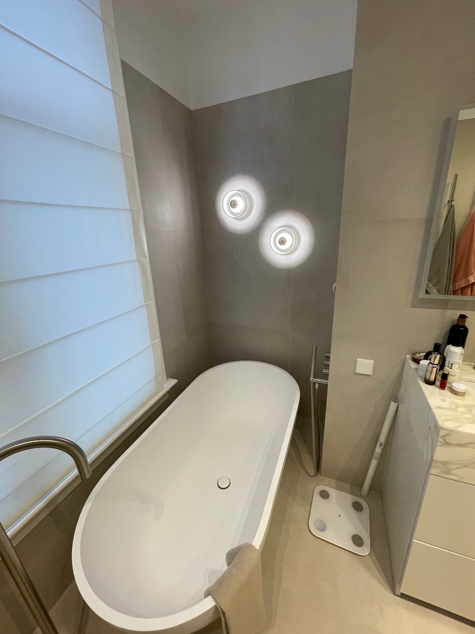 Nowoczesna łazienka w beżowych tonacjach z wolnostojącą wanną, okrągłymi lampami na ścianie i roletami rzymskimi w oknie. Minimalistyczny design i eleganckie wykończenie.