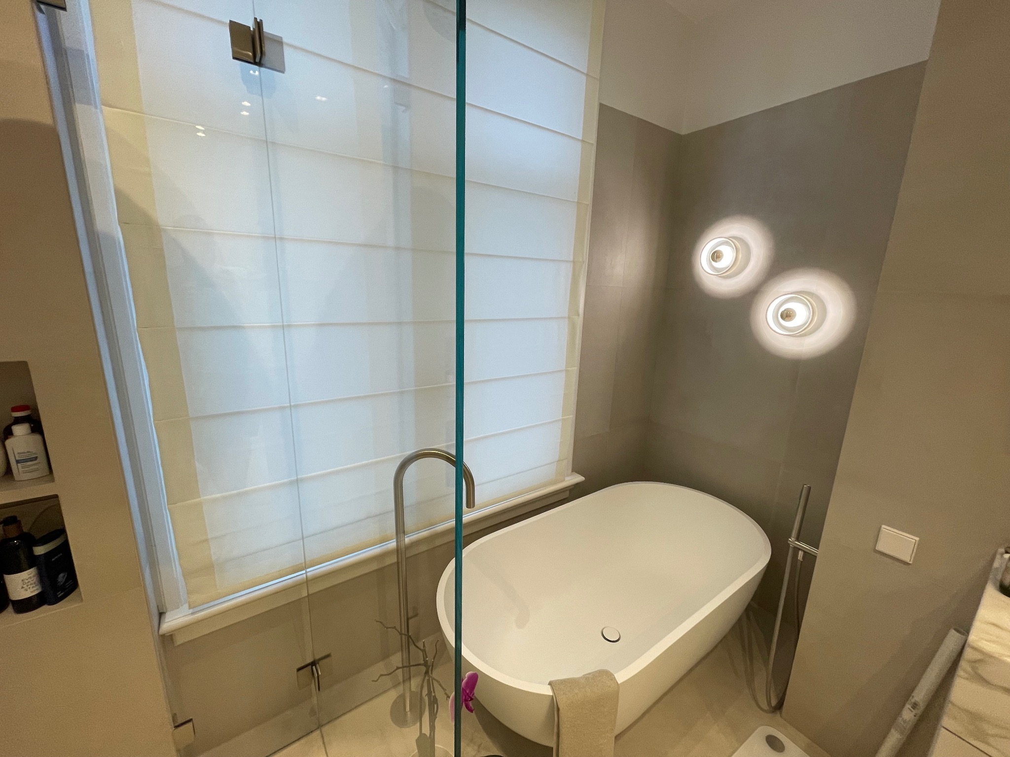 Nowoczesna łazienka z wolnostojącą wanną, szklaną kabiną prysznicową i roletami rzymskimi. Dwa okrągłe, dekoracyjne kinkiety oświetlają ścianę.