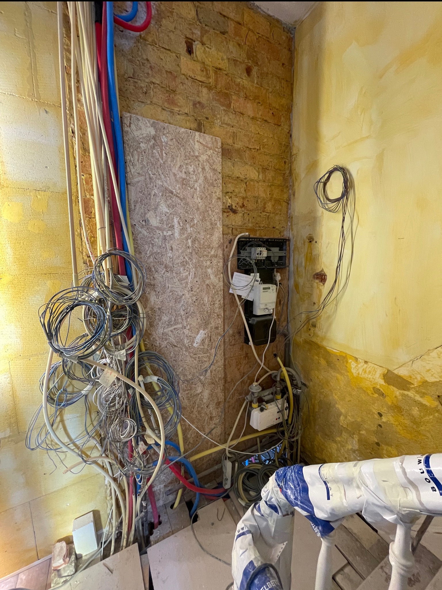 Niezabezpieczone kable i rury w trakcie prac remontowych w pomieszczeniu z odsłoniętą cegłą i żółtą ścianą. Widoczna skrzynka z licznikiem.