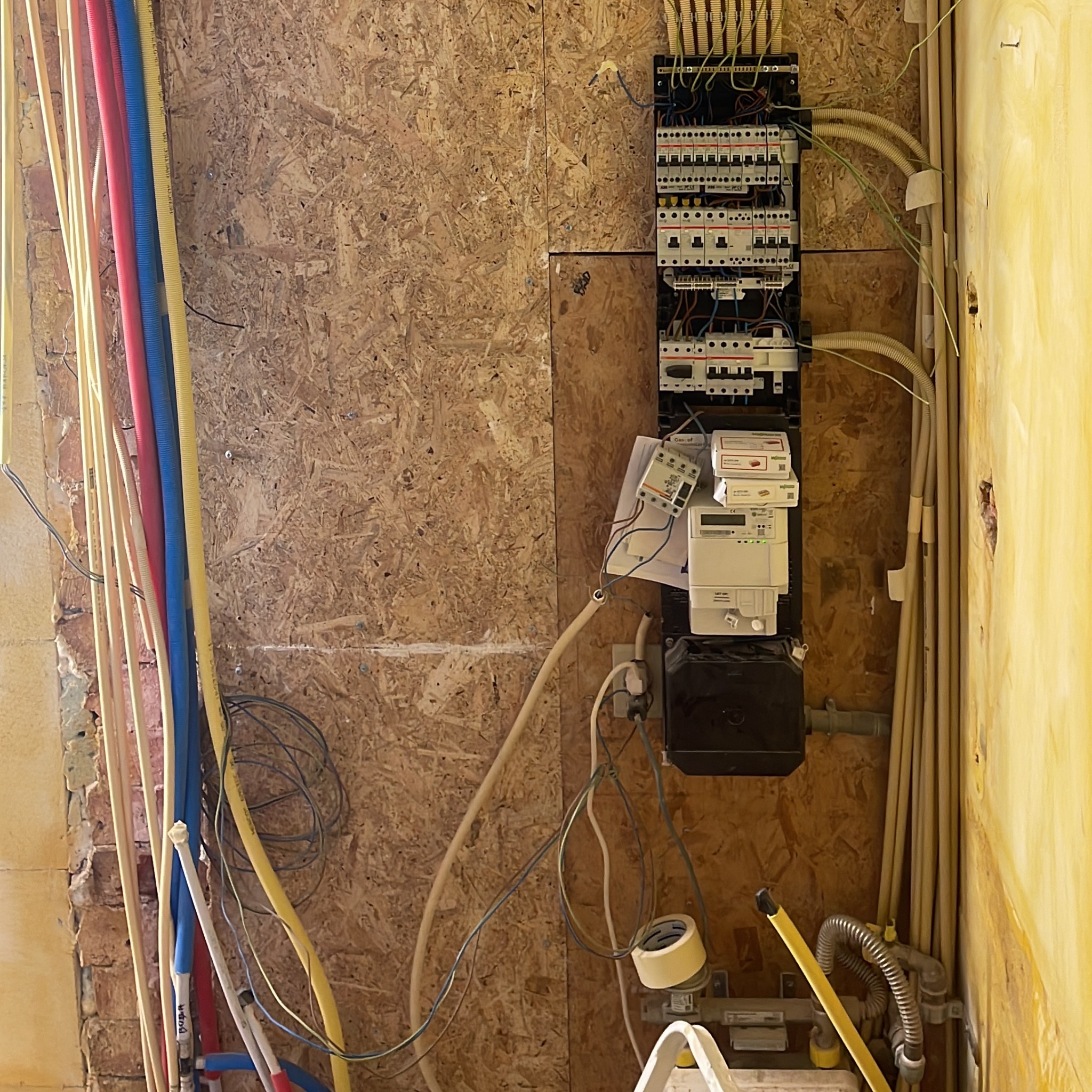 Rozdzielnia elektryczna z licznikami na ścianie z płyty OSB, obok kolorowe rury instalacyjne. Widoczne przewody i elementy montażowe. Prace w trakcie realizacji.