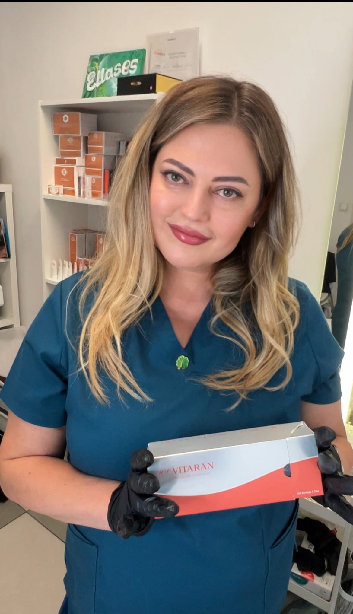 Kobieta w uniformie medycznym prezentuje opakowanie Vitaran, preparatu do biorewitalizacji skóry. Widoczny fragment regału z kosmetykami i akcesoriami.