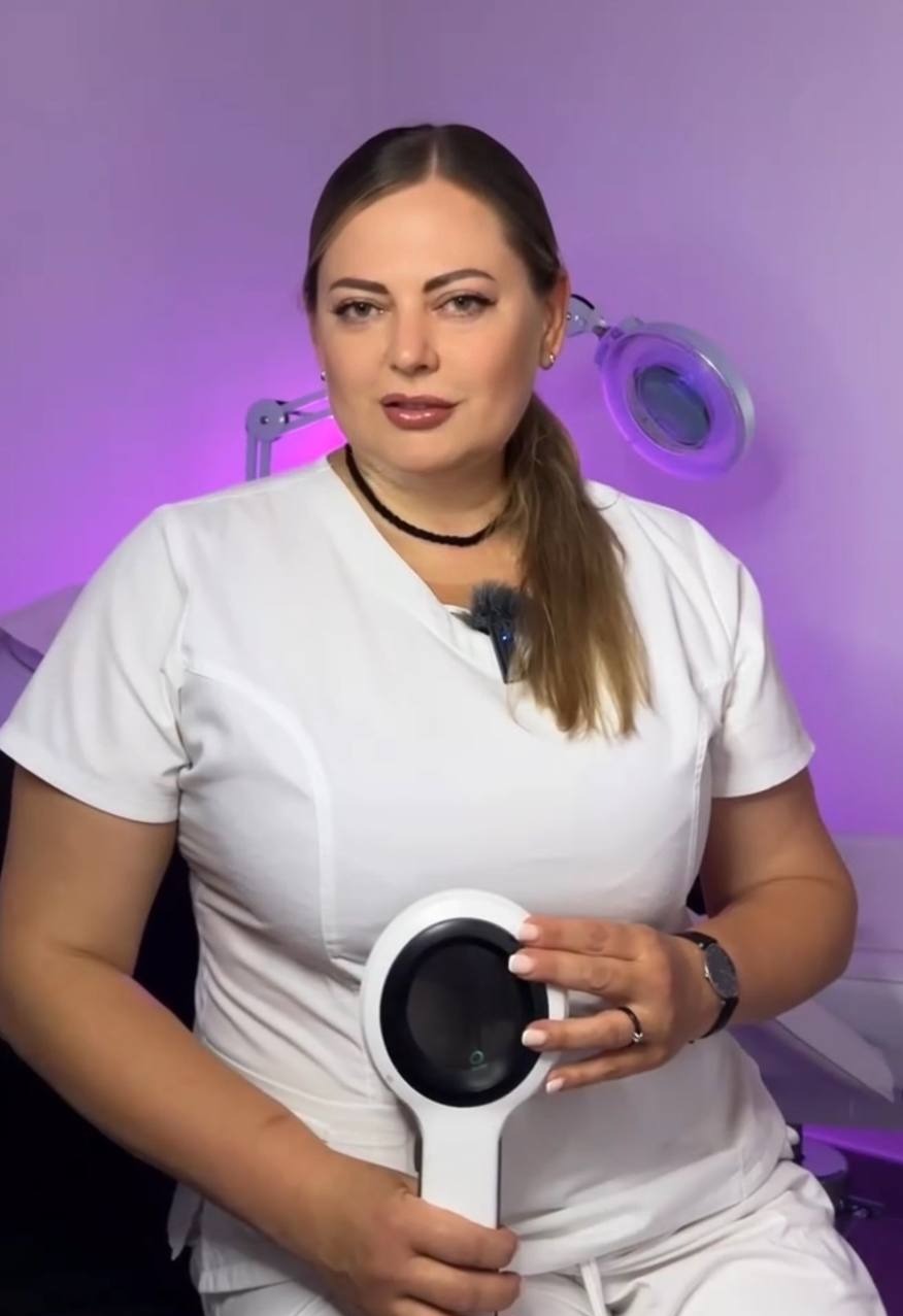 Kobieta w białym uniformie prezentuje dermatoskop na tle fioletowego oświetlenia, sugerując wizytę dermokosmetologiczną w salonie.