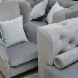 Szary fotel z poduszką i sofa z guzikami, obite miękkim welurem. Fotel zdobiony nitami. Całość w chłodnej tonacji szarości.