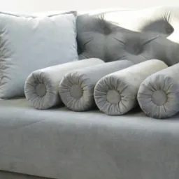 Szary, welurowy narożnik z poduchami i wałkami dekoracyjnymi, widoczne pikowania i detale wykończenia. Stonowana kolorystyka, miękkie światło.