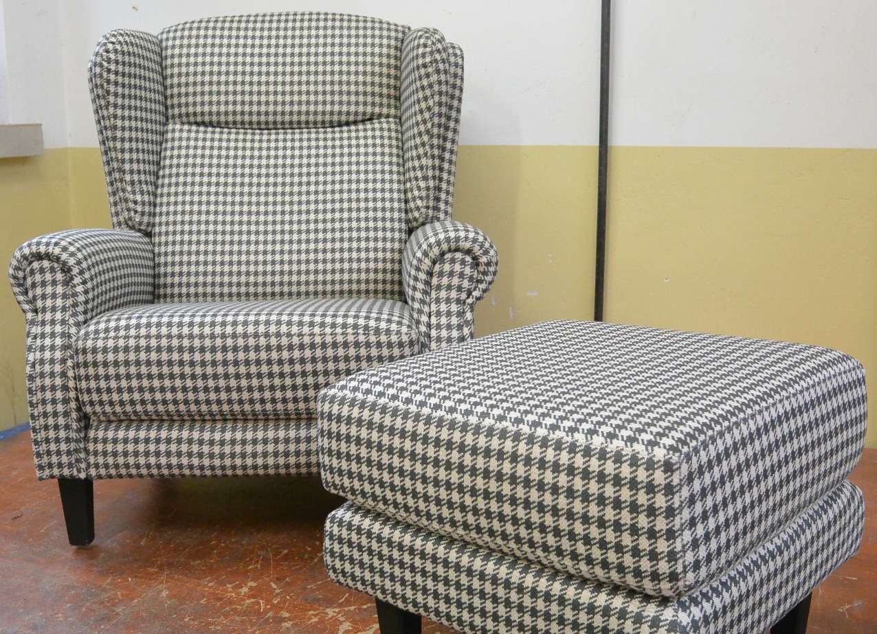 Fotel typu uszak w pepitkę z podnóżkiem, na tle dwukolorowej ściany. Klasyczny design, widoczne staranne tapicerowanie i dbałość o detale.