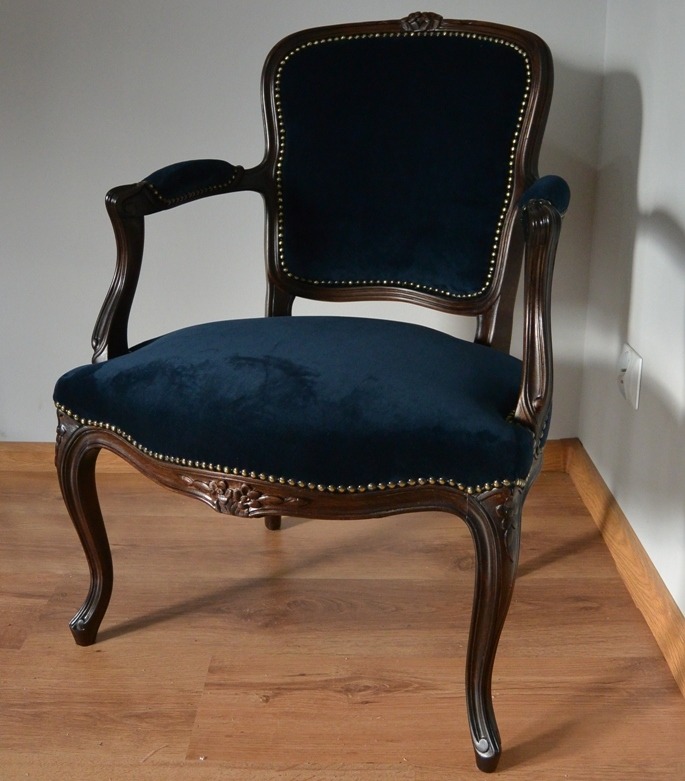Stylowy, granatowy fotel tapicerowany z ciemnego drewna, wykończony ozdobnymi ćwiekami, na drewnianej podłodze. Widok z przodu, lekko z ukosa.