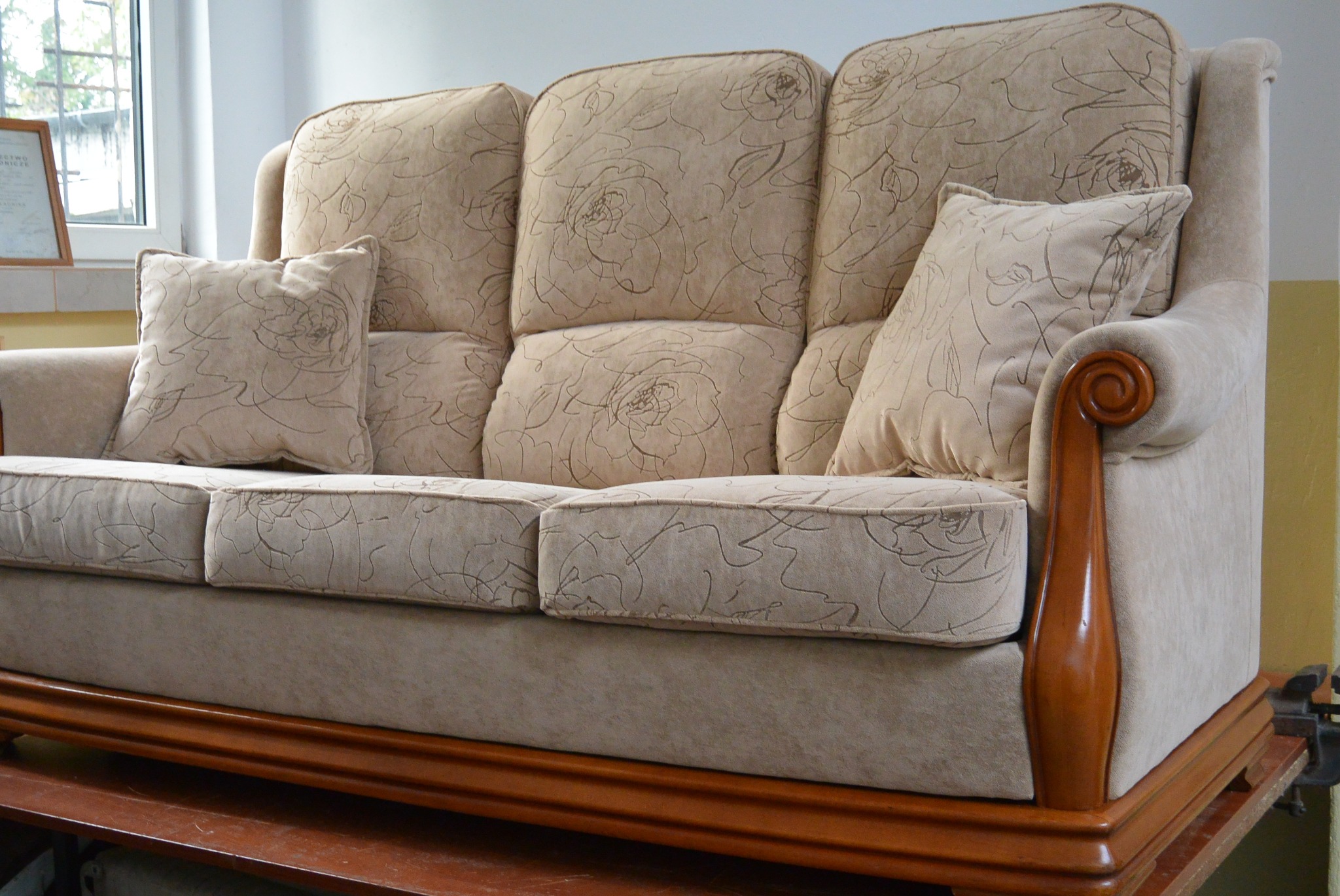 Beżowa, tapicerowana sofa trzyosobowa z poduszkami w kwiatowe wzory, na drewnianej podstawie. Widoczny fragment okna i dokument w ramce w tle.