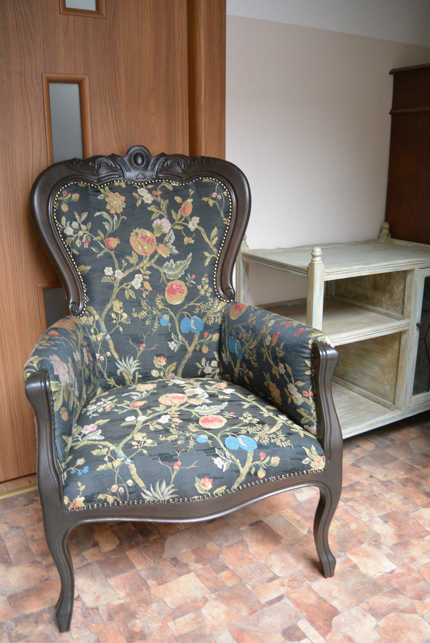 Stylowy, tapicerowany fotel z ciemną drewnianą ramą i wzorzystą tkaniną w kwiaty. Obok stolik z półkami, brązowa podłoga i drzwi.