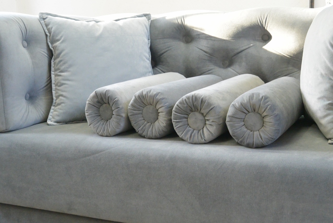 Szary, welurowy narożnik z poduchami i wałkami dekoracyjnymi, widoczne pikowania i detale wykończenia. Stonowana kolorystyka, miękkie światło.