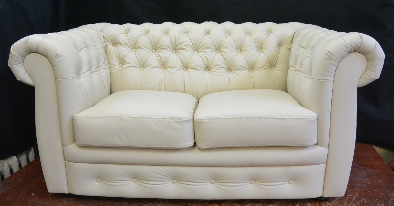 Elegancka, dwuosobowa sofa w stylu Chesterfield, obita jasną skórą ekologiczną, z charakterystycznymi pikowaniami i poduszkami siedziskowymi.