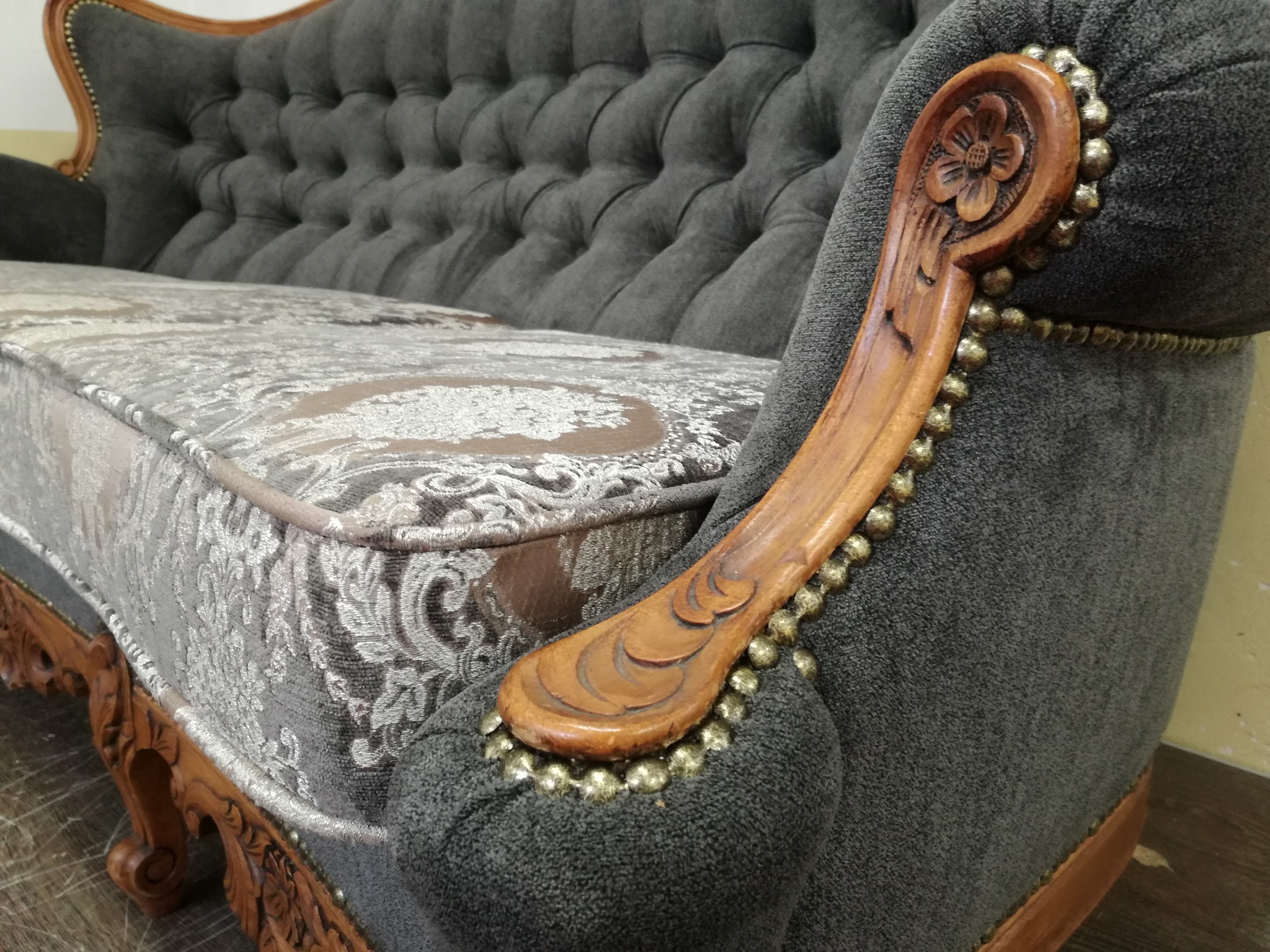 Szary, tapicerowany fotel z drewnianymi zdobieniami i wzorzystą poduszką siedziska. Detale wykończenia z nitami. Widok z boku, ukazujący rzeźbiony podłokietnik.